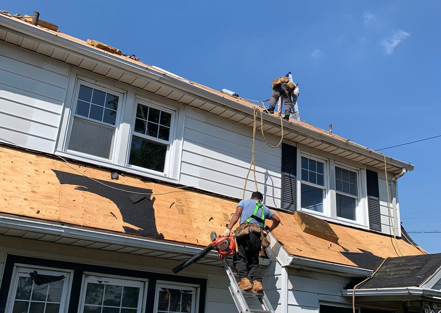 Two workers inspecting and repairing the roof of a residential house using safety ropes and ladders.