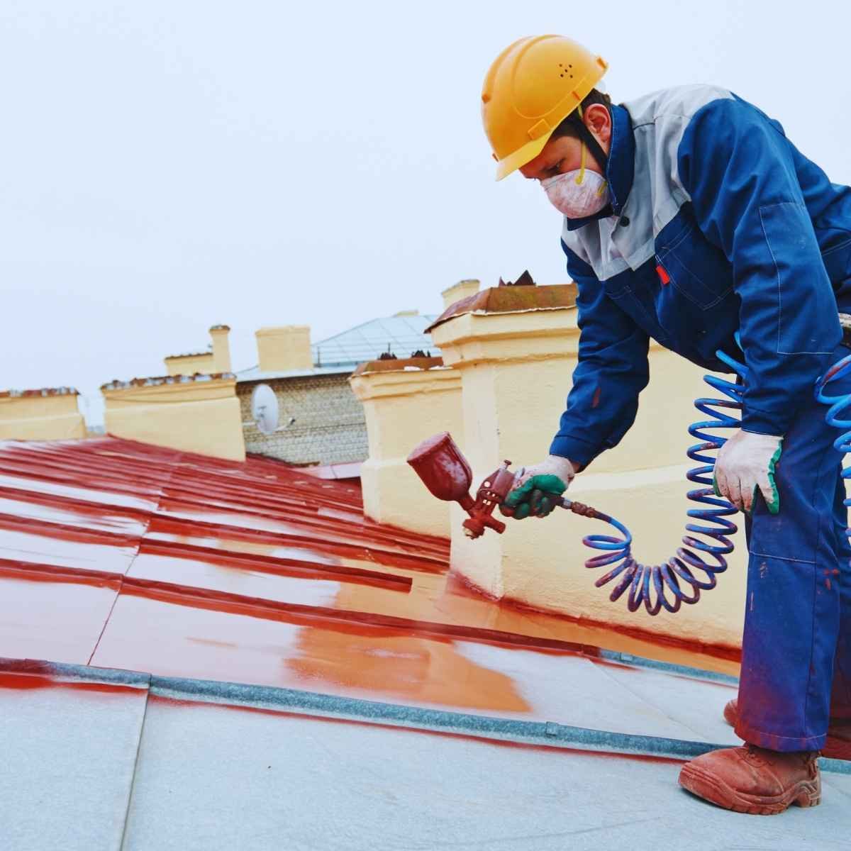 Roofer in safety gear applying red acrylic coating on a sloped metal roof using a spray gun.